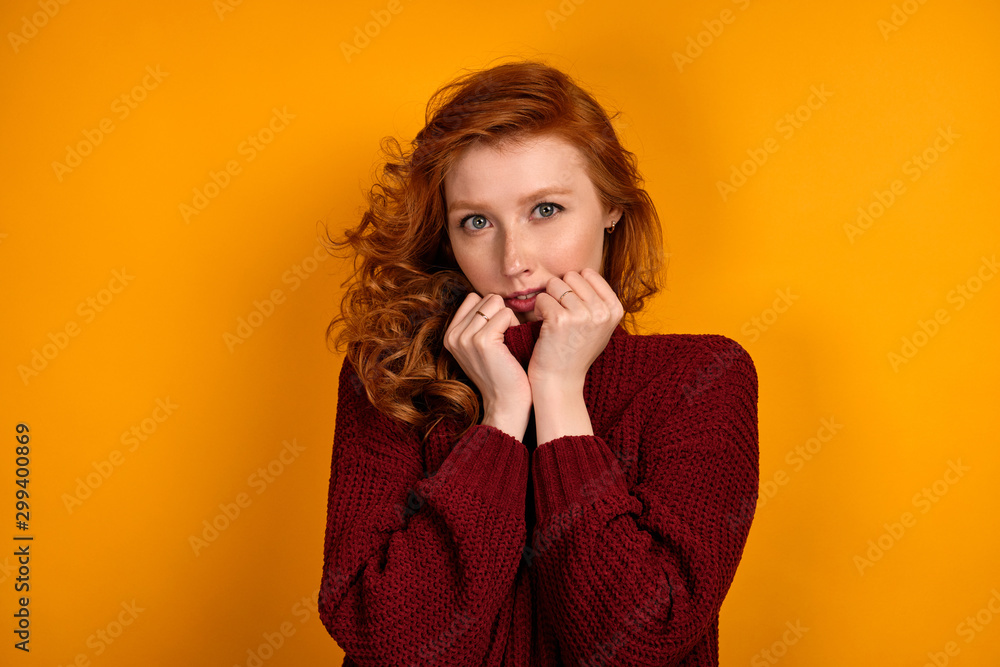 Redhead girl with a shock of curls in a burgundy sweater stands on a yellow background, raising the collar of a sweater. Redhead girl with a shock of curls in a burgundy sweater stands on a yellow background, raising the collar of a sweater.