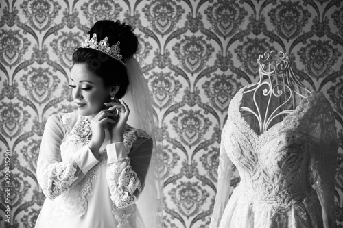Bride portrait face closeup sensual make up and diadem on her head, corrects earrings. Beautiful charming bride in white robe standing near the mannequin with wedding dress in the room in the morning © Tetiana Moish