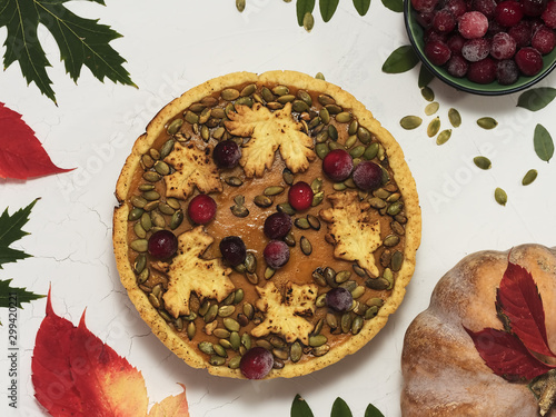 Homemade American pumpkin pie on a white plate decorated with pumpkin seeds and cranberries. Against the background of autumn leaves, pumpkin seeds, ripe pumpkin and a cup with cranberries. Top view.
