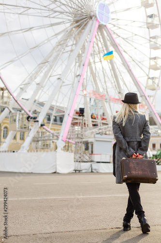 Back view of female wearing hat coat walking away . She carrying old suitcase over wheel background