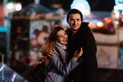 Photo of a smiling loving couple walking outdoors in amusement park having fun hugging in the evening