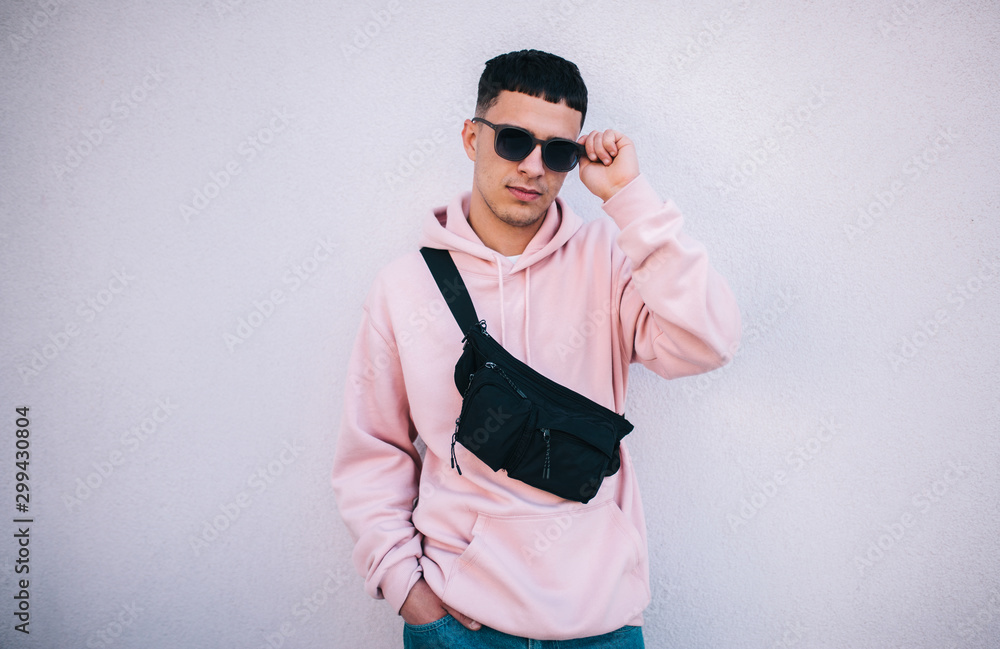 City portrait of handsome hipster guy wearing pink blank hoodie with waist black bag with space for your logo or design. Mockup for print City portrait of handsome hipster guy wearing pink blank hoodie with waist black bag with space for your logo or design. Mockup for print