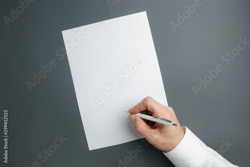 Male hand holding pen and writing on empty sheet on grey background for text or design. Blank templates for contact, advertising or use in business. Finance, office, purchases. Mock up. Copyspace.