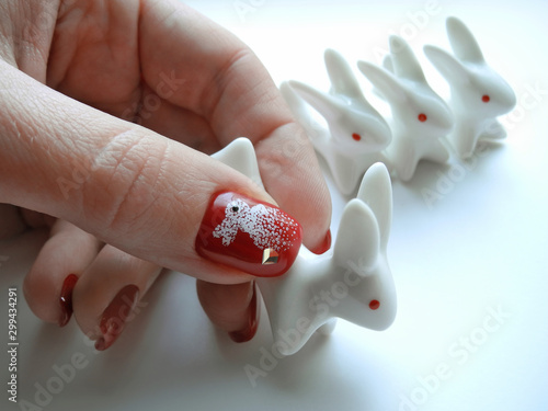 Female soigne hand with bright festive red manicure holds white porcelain rabbit on background of number of the same statuettes. photo