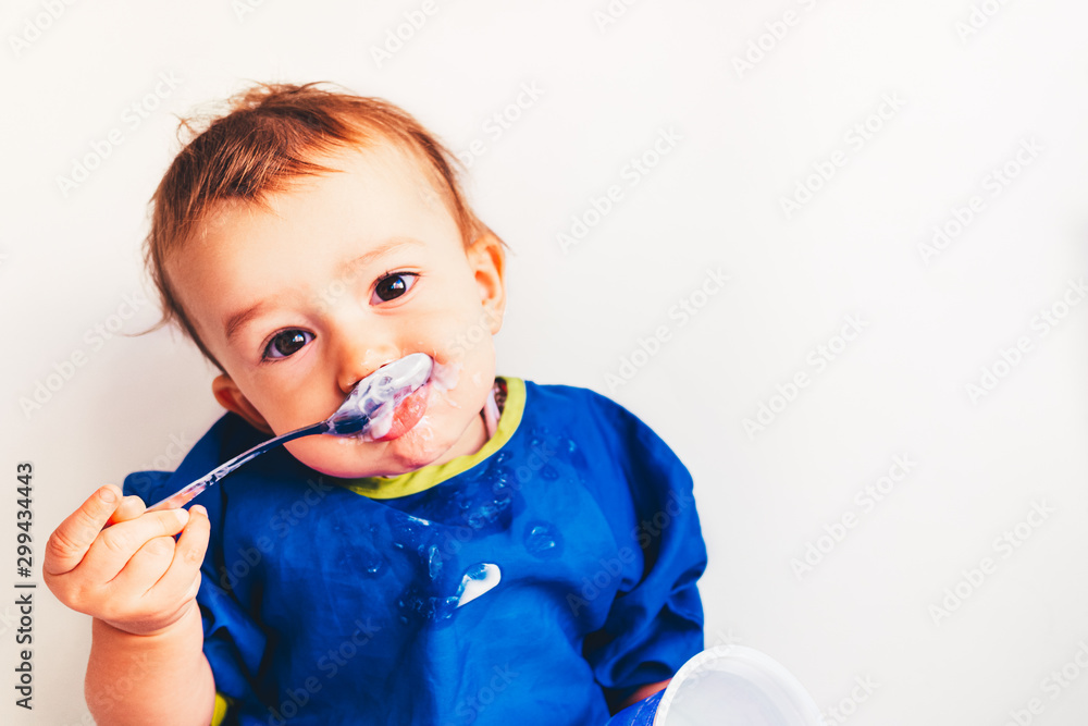 Baby savoring his first yogurt with a spoon, with a spotted face and adorable expression. - obrazy, fototapety, plakaty 