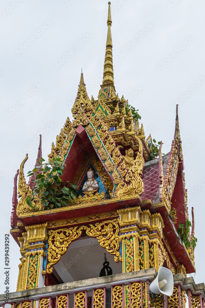 Fototapeta premium Ko Samui Island, Thailand - March 18, 2019: Wat Laem Suwannaram Chinese Buddhist Temple. Tower and spire of small golden shrine with statue, loudspeaker, blues and green foliage against silver sky. 