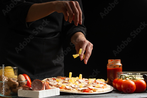 Chef cooks Italian pizza  sprinkles with pineapple. Freezing in motion. Against the background of pizza ingredients. Black background with space for design.