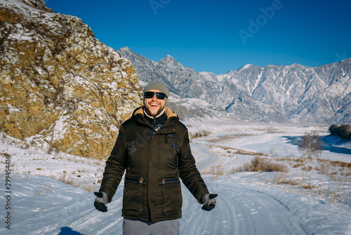 Young cheerful guy enjoying Christmas vacation in mountains. Attractive man laughs and gives thumbs up against snow-capped mountains and blue sky. New year's journey, active winter tourism concept.