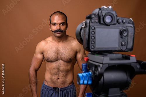 Portrait of muscular Indian man with mustache shirtless vlogging with camera