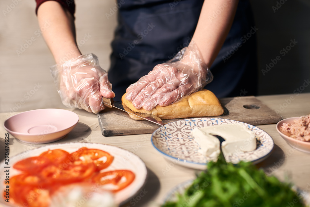 Focus on hand cutting with knife long bread roll on cutting board for preparing grilled panini sandwich. Ingredients on blurred foreground: tomato slices, tuna, butter, salad on plate at wooden table.