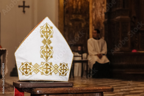 Mitra, copricapo religione Cattolica, appoggiato su un banco durante una cerimonia, chierico sullo sfondo seduto. photo