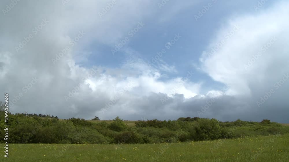 Wide shot of a flat grassy field, behind which is a tree-dotted hill, beneath a sky changing from blue to being heavy with clouds Wide shot of a flat grassy field, behind which is a tree-dotted hill, beneath a sky changing from blue to being heavy with clouds