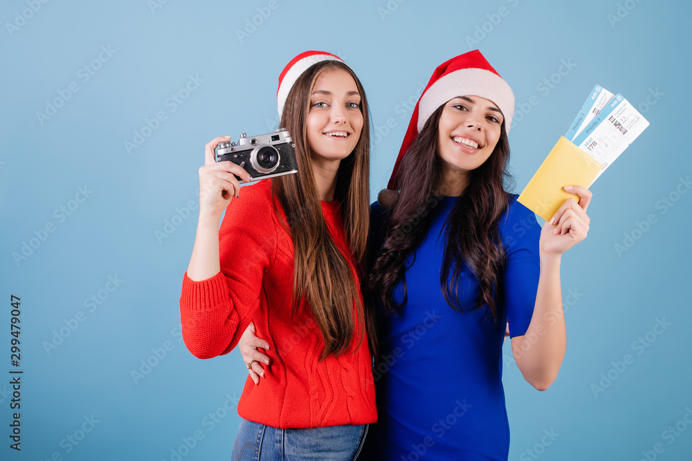 two women wearing santa hats with plane tickets and passport and vintage camera isolated over blue two women wearing santa hats with plane tickets and passport and vintage camera isolated over blue