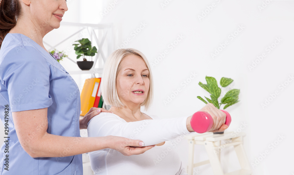 Modern rehabilitation physiotherapy. Physiotherapist helps an middle aged woman recover from an injury through exercise with dumbbells. Modern rehabilitation physiotherapy. Physiotherapist helps an middle aged woman recover from an injury through exercise with dumbbells.