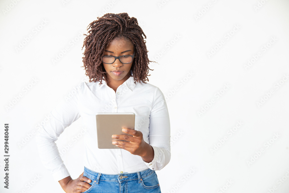 Focused serious expert in glasses using tablet, reading on screen. Young African American female professional standing isolated over white background. Digital communication concept Focused serious expert in glasses using tablet, reading on screen. Young African American female professional standing isolated over white background. Digital communication concept