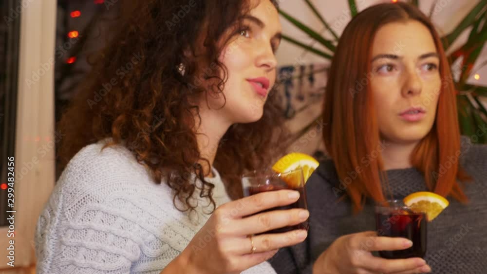 Girls with a glass of mulled wine communicate at a festive party. Happy friends together. Two young women celebrating new years eve party at home. Holidays and celebration concept. 4K footage Close-up Girls with a glass of mulled wine communicate at a festive party. Happy friends together. Two young women celebrating new years eve party at home. Holidays and celebration concept. 4K footage Close-up