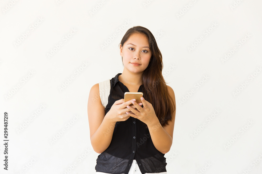 Woman using smartphone and looking at camera. Beautiful focused young female student with backpack standing and messaging via cell phone on white background. Technology concept Woman using smartphone and looking at camera. Beautiful focused young female student with backpack standing and messaging via cell phone on white background. Technology concept