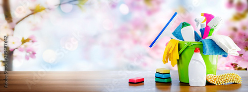 Bucket or basket with cleaning items on wooden table and blurry pink flowers tree background. Washing set with copy space banner.