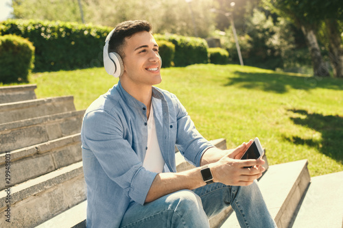 Outdoors Leisure. Stylish guy in headphones wearing watch sitting on stairs in park with smartphone listening to music smiling happy