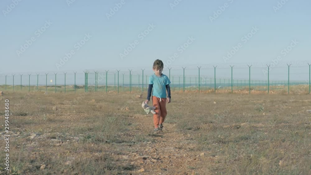 one lonely poor dirty little boy with toy walking along desert near state border with fence and razor barbed wire head down one lonely poor dirty little boy with toy walking along desert near state border with fence and razor barbed wire head down