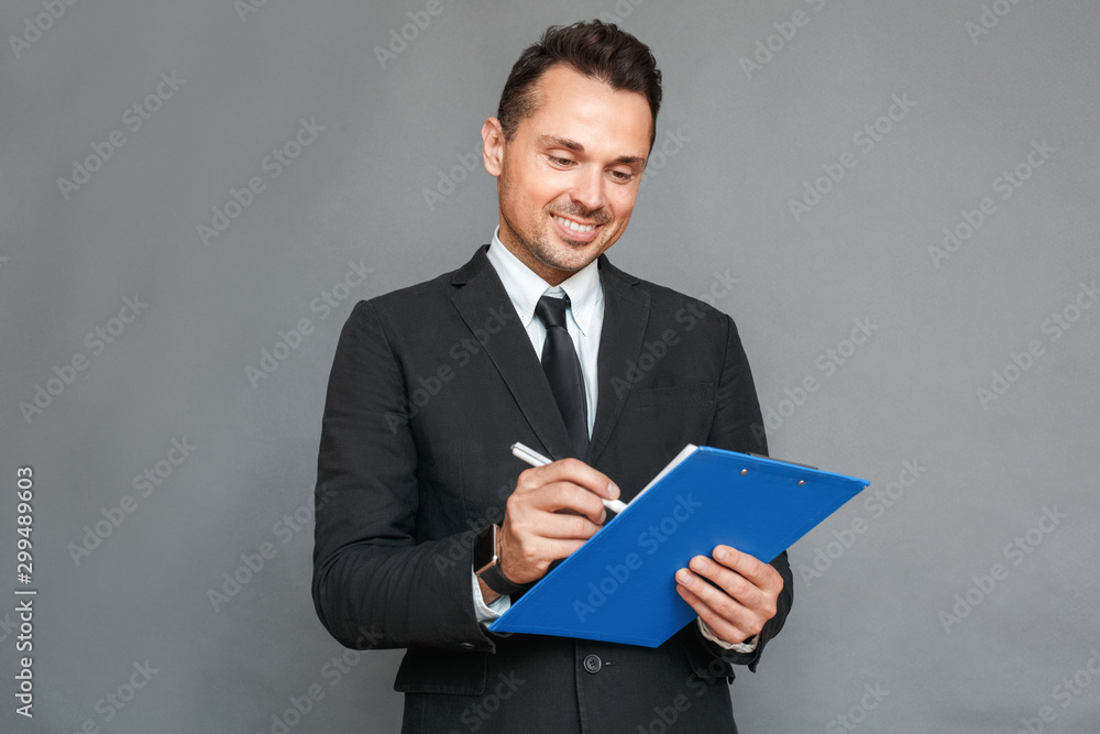 Businessman studio standing isolated on grey with clipboard taking notes smiling joyful Businessman studio standing isolated on grey with clipboard taking notes smiling joyful