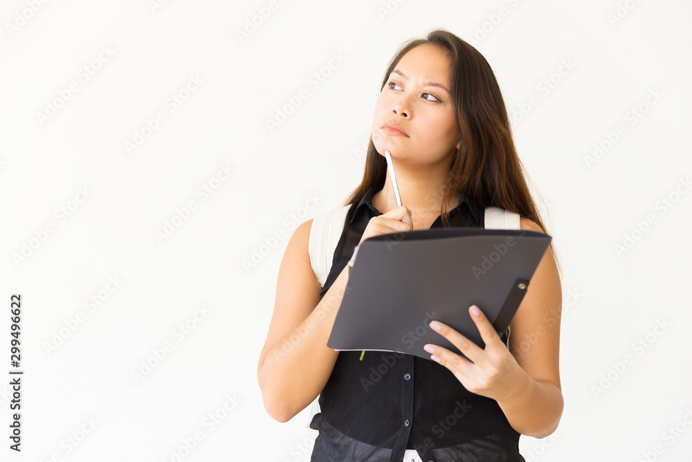 Thoughtful young woman with folder and pen. Pensive young female student with backpack holding folder with pen and looking away. Education concept Thoughtful young woman with folder and pen. Pensive young female student with backpack holding folder with pen and looking away. Education concept