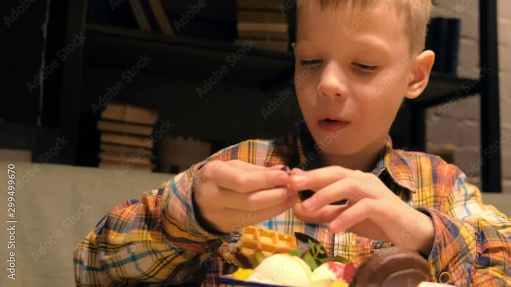 Portrait of child boy is eating sweet tasty dessert with ice cream, cookies, waffles and fruit in white plate in cafe. Enjoying the food. Unhealthy dessert. Portrait of child boy is eating sweet tasty dessert with ice cream, cookies, waffles and fruit in white plate in cafe. Enjoying the food. Unhealthy dessert.