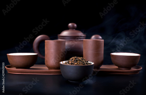 Asian tea ceremony. Clay teapot and tea cups with vapor on a black background.