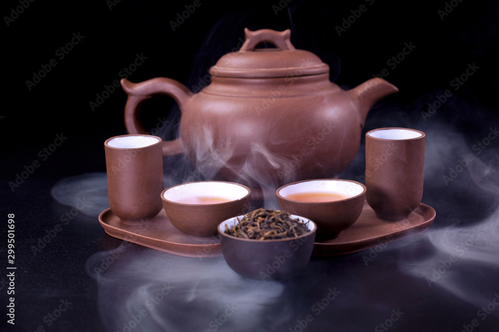 Asian tea ceremony. Clay teapot and tea cups with vapor on black background.