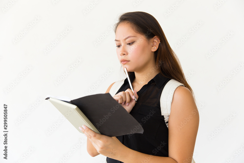 Serious focused student girl studying her notes, holding pen, reading open notebook. Young Latin woman standing isolated over white background. Studying concept Serious focused student girl studying her notes, holding pen, reading open notebook. Young Latin woman standing isolated over white background. Studying concept