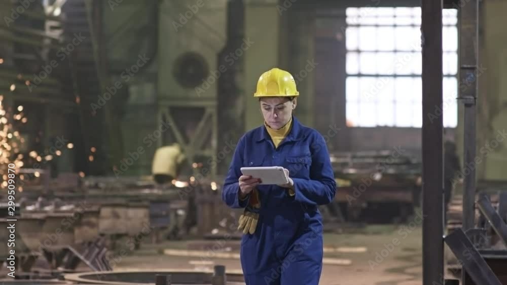 Tracking shot of busy female engineer in hard hat working on tablet and walking through metal fabrication facility Tracking shot of busy female engineer in hard hat working on tablet and walking through metal fabrication facility