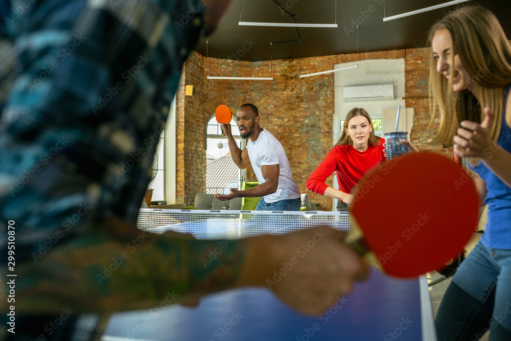 Obraz premium Young people playing table tennis in workplace, having fun. Friends in casual clothes play ping pong together at sunny day. Concept of leisure activity, sport, friendship, teambuilding, teamwork.
