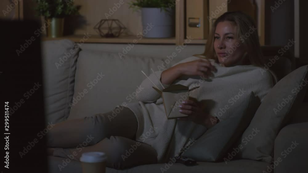 Dolly-in shot of woman relaxing on couch in dark living room and eating Chinese takeaway while watching TV Dolly-in shot of woman relaxing on couch in dark living room and eating Chinese takeaway while watching TV