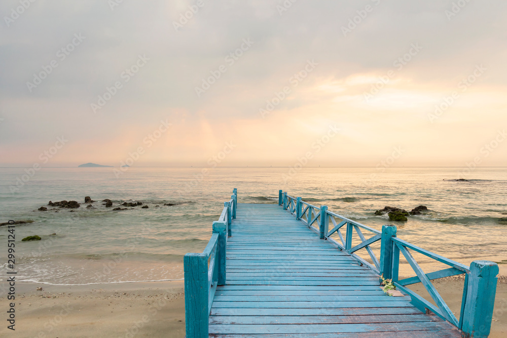 Wood bridge blue on the sea which has walk way for travel tourism and beautiful sky with sunshine in the morning. Sea waves slowly a the splashing on the sand and rocks with moss background.