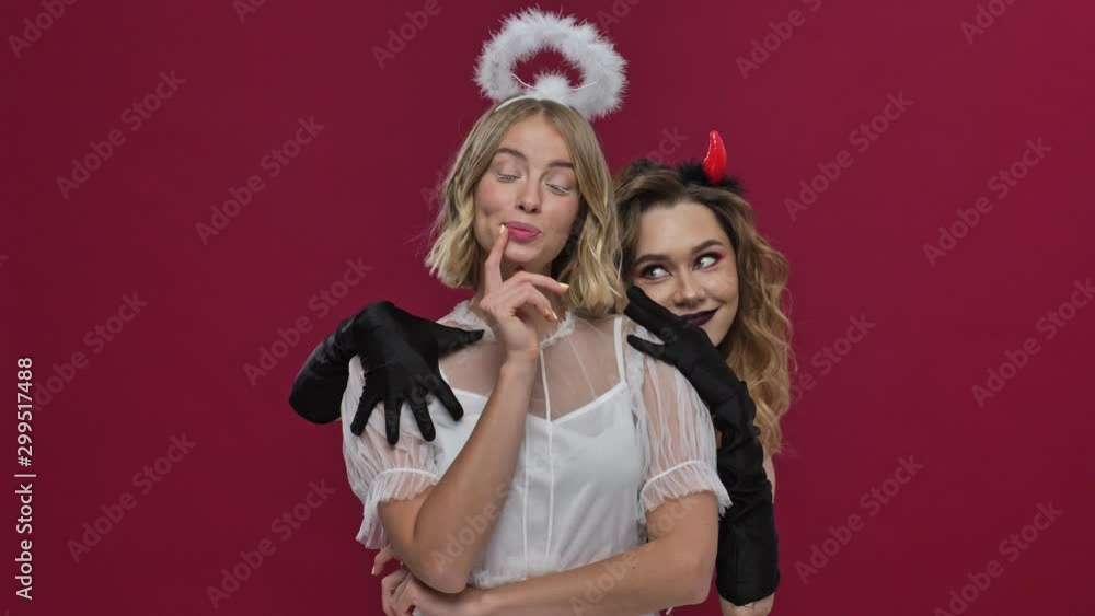 Hungry demon girl hugs from behind a cute angel girl in carnival costumes isolated over red wall background Hungry demon girl hugs from behind a cute angel girl in carnival costumes isolated over red wall background
