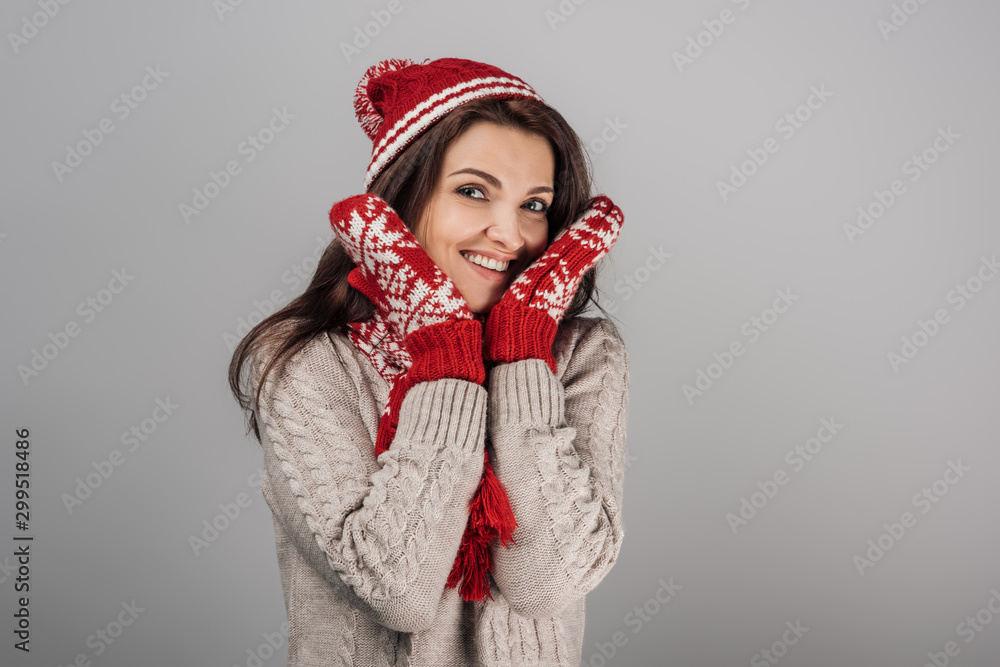 cheerful woman in gloves and knitted hat touching face isolated on grey