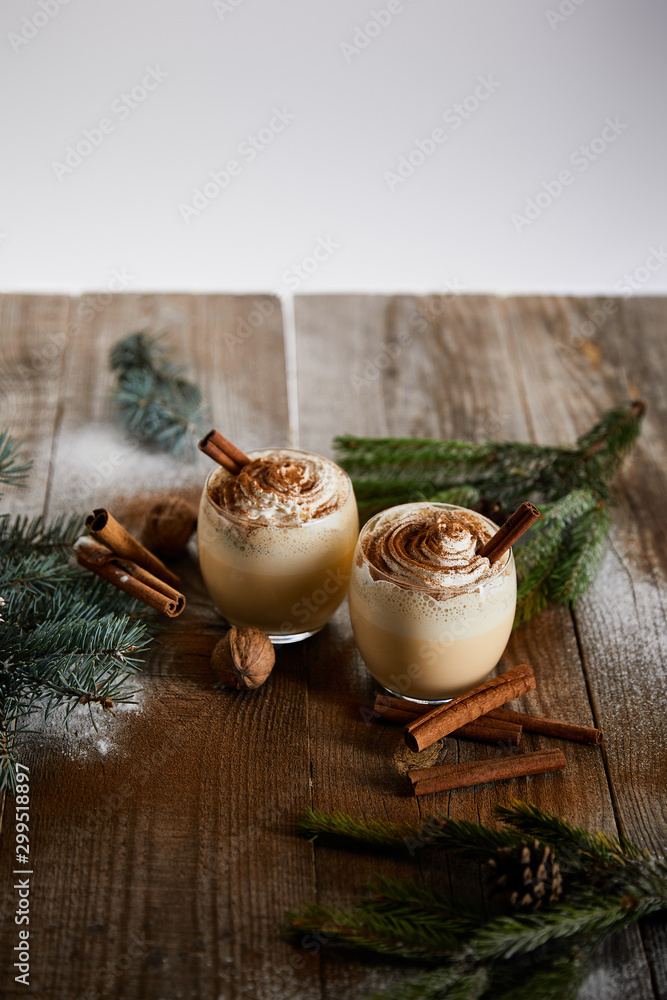 eggnog cocktail with whipped cream and cinnamon near spruce branches and powdered sugar scattered on wooden table isolated on grey - obrazy, fototapety, plakaty 