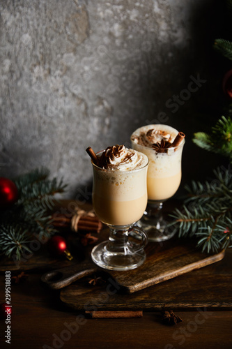 delicious eggnog cocktail with whipped cream and cinnamon on cutting board near spruce branches on grey stone background