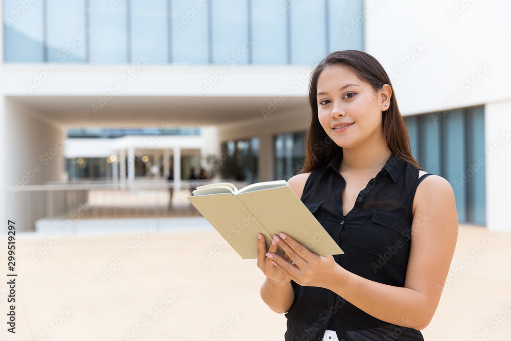 Positive female student reading book outside. Young Latin woman standing near college building, holding open notebook, looking at camera. Knowledge concept Positive female student reading book outside. Young Latin woman standing near college building, holding open notebook, looking at camera. Knowledge concept