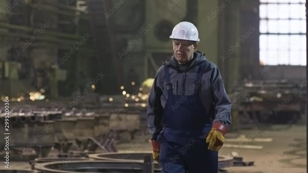 Tracking shot of tired elderly male worker in overalls, gloves and hard hat walking through metal fabrication facility Tracking shot of tired elderly male worker in overalls, gloves and hard hat walking through metal fabrication facility