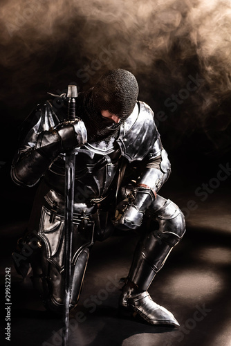 handsome knight in armor holding sword and bend knee on black background © LIGHTFIELD STUDIOS