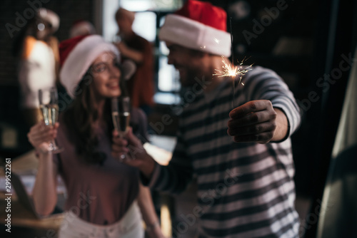selective focus of happy businessman and businesswoman in santa hats looking at each other while holding champagne glasses