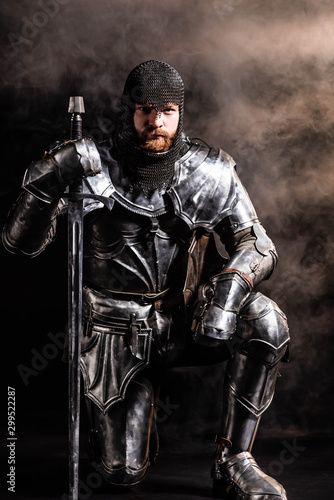 handsome knight in armor holding sword and bend knee on black background © LIGHTFIELD STUDIOS