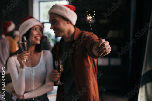 selective focus of young businessman holding sparkler near attractive, smiling colleague