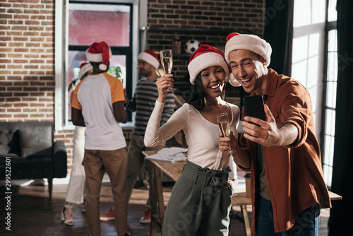 cheerful businessman and businesswoman in santa hats taking selfie on smartphone while holding champagne glasses