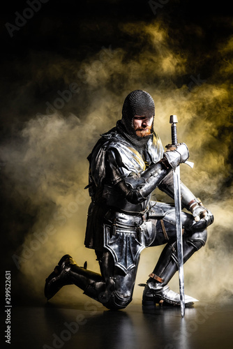 Foto handsome knight in armor holding sword and bend knee on black background