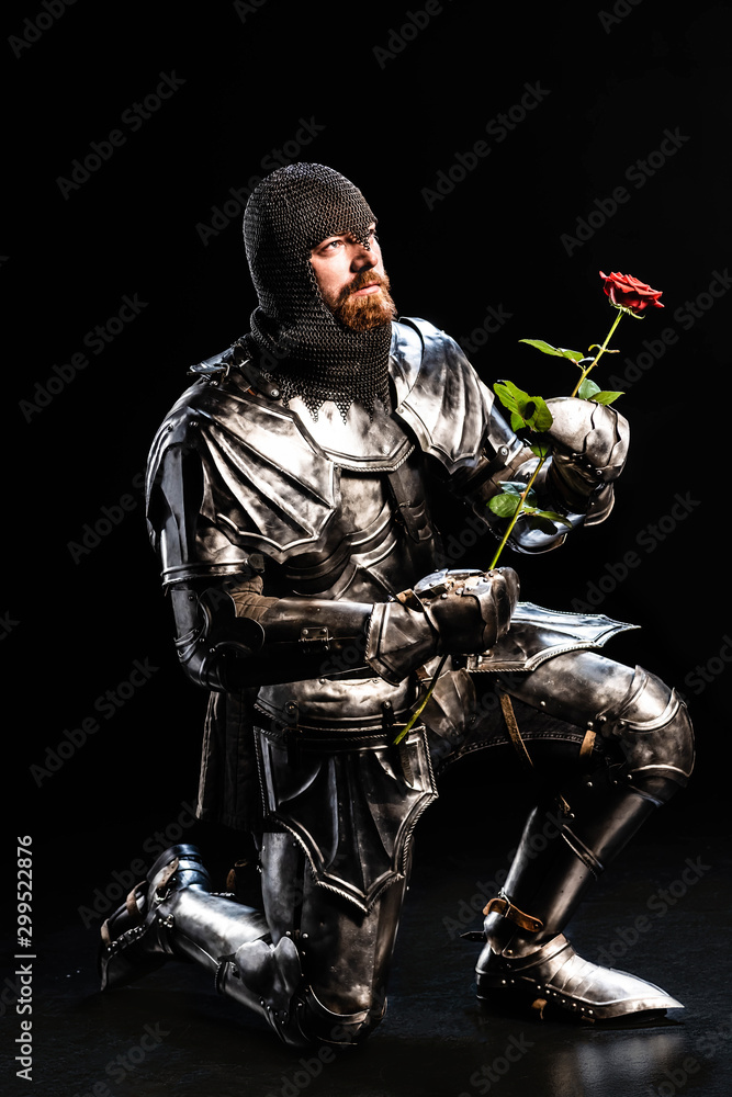 Naklejka premium handsome knight in armor holding rose and bend knee on black background