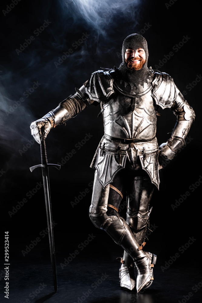 Naklejka premium smiling knight in armor looking at camera and holding sword on black background