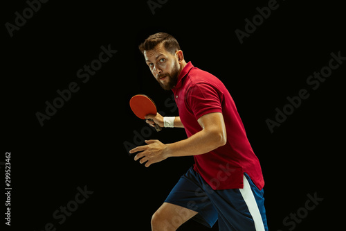 Young man plays table tennis on black studio background. Model in sportwear plays ping pong. Concept of leisure activity, sport, human emotions in gameplay, healthy lifestyle, motion, action, movement photo