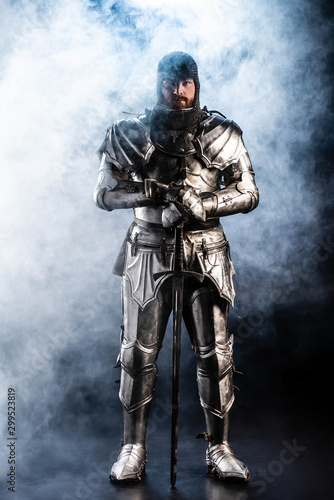 handsome knight in armor looking at camera and holding sword on black background